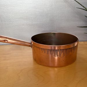 May include: A copper saucepan with a long handle. The pot has a cylindrical shape with a decorative textured pattern around the base. The handle is attached to the side of the pot. The pot is sitting on a wooden surface.