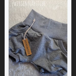 May include: Gray cotton baby shorts with a striped waistband and a brown tag that says "HANDMADE". The shorts have a pocket on the back. The text "ZWERGENNÄHSTUBE" is visible at the top of the image.