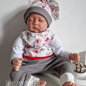 Könnte beinhalten: Eine Babypuppe trägt eine graue und weiße Mütze mit Blumenmuster, eine Lätzchen und eine Hose. Das Outfit hat rote Verzierungen. Die Puppe sitzt auf einer weißen Oberfläche mit dem Text "ZWERGENNÄHSTUBE" sichtbar.