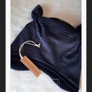 Op de afbeelding: Een donkerblauwe geribbelde gebreide muts met een fleecevoering en een geknoopte bovenkant. De muts heeft een bruin label met de tekst "Handmade" en "Zwergennahstube".