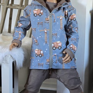 Könnte beinhalten: Ein kleines Kind trägt eine blaue Kapuzenjacke mit einem Baufahrzeug-Druck, eine braune Hose und eine braune Mütze. Das Kind steht auf einem Holzboden.