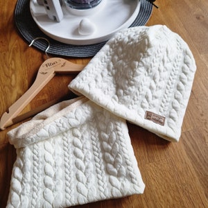 Könnte beinhalten: Zwei weiße Strickwaren, eine Mütze und ein Halstuch, mit einem Zopfmuster. Die Mütze hat ein kleines braunes Lederlabel mit dem Text "Zwergen" und "Trachtenmode".