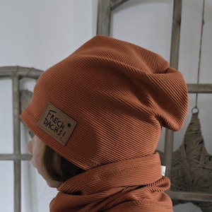 Puede incluir: Conjunto de gorro y braga de cuello de punto acanalado color óxido. El gorro presenta un pequeño parche de cuero con el texto "FRECH DACHS!". La braga de cuello está envuelta alrededor del cuello. El conjunto es de tejido suave y texturizado.