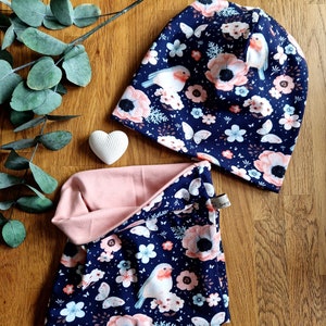 Op de afbeelding: Een blauwe en roze gepatroneerde muts en nekwarmer set met een bloemen- en vogelontwerp. De muts heeft een omslagmanchet en de nekwarmer is omkeerbaar met een roze kant.