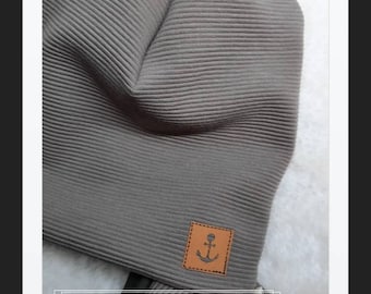 以下が含まれることがあります： 黒い錨のデザインが施された茶色の革パッチが付いた、グレーのリブ編みビーニー。テキスト「FERN DER STADT」は、画像の下部にある白いボックスに表示されます。