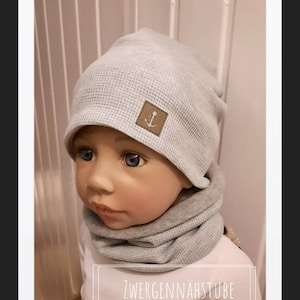 Puede incluir: Conjunto de gorro y braga de cuello de punto gofrado gris claro. El gorro presenta un pequeño parche marrón con una ancla bordada. El texto "ZWERGENNÄHSTUBE" se muestra en un recuadro blanco en la parte inferior de la imagen.