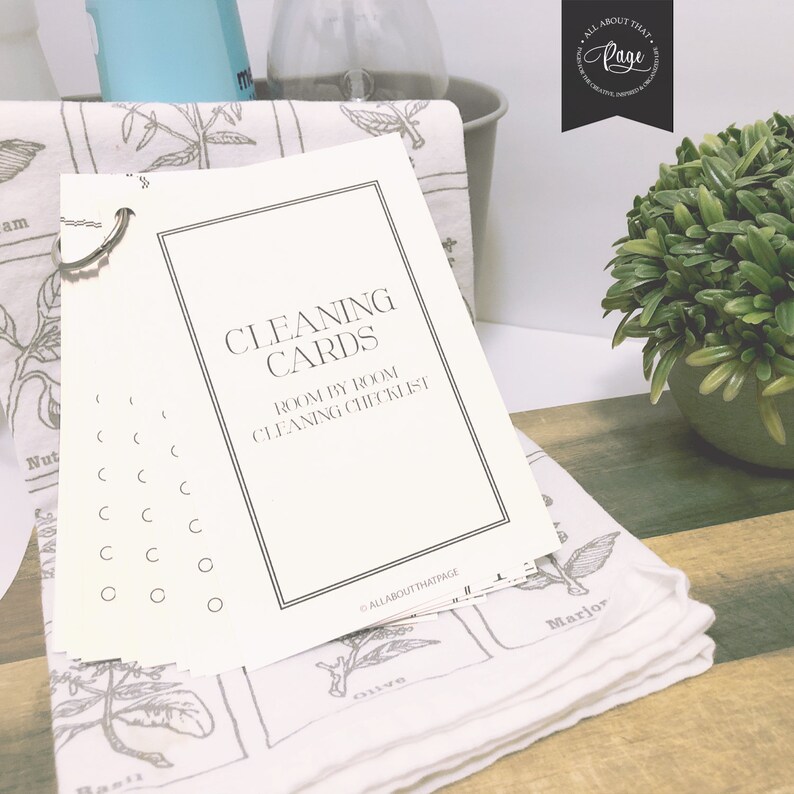 May include: A set of printable cleaning cards with the text "Cleaning Cards Room by Room Cleaning Checklist" on a white background. The cards are on a white towel with a botanical print.