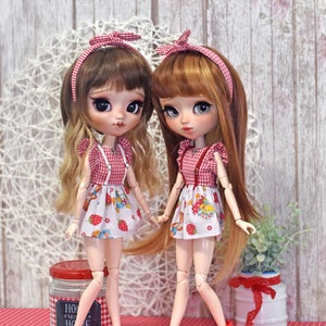 Könnte beinhalten: Zwei Puppen mit langen Haaren, die passende rot-weiß karierte Kleider und Stirnbänder tragen. Die Kleider haben einen Erdbeer- und Fruchtdruck. Eine Puppe trägt rote Ballerinas, die andere rote Stiefel. Die Puppen stehen auf einer roten Oberfläche.