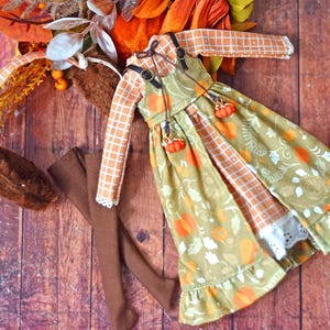 Op de afbeelding: Een herfstoutfit voor poppen met een oranje geruit shirt met lange mouwen, een groene jurk met pompoen- en bladerpatronen en bruine leggings. De jurk heeft pompoenbedels en witte kanten afwerking. Een herfsthoofdband is ook aanwezig.