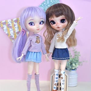 Può includere: Due bambole alla moda con tratti del viso dettagliati e capelli lunghi. Una bambola ha i capelli viola e indossa una maglietta a righe, una gonna blu e scarpe da ginnastica viola. L'altra bambola ha i capelli castani e indossa un outfit simile con una gonna di jeans e stivali marroni.