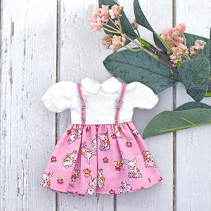 Può includere: Un vestito per bambole con corpetto bianco con colletto Peter Pan e maniche a sbuffo, abbinato a una gonna rosa ornata da una stampa di coniglietti e fiori. L'abito ha bretelle rosa ed è ambientato su uno sfondo di legno bianco con accenti floreali.