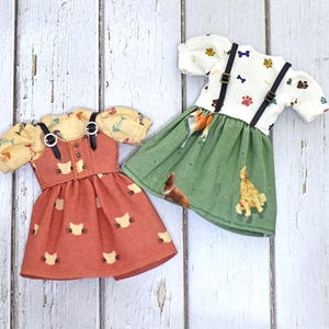 May include: Two doll dresses. One is orange with cat faces, the other is white with paw prints and dog bone designs. Both have suspenders and short sleeves. The dresses are on a white wooden surface.