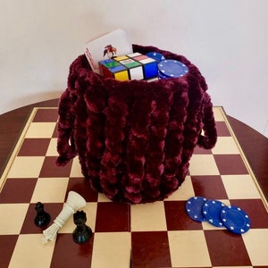 Pode incluir: Uma bolsa de dados de pelúcia bordô com fecho de cordão, cheia de um cubo de Rubik, fichas de pôquer e um cartão de jogo. A bolsa está sobre um tabuleiro de xadrez de madeira com peças de xadrez.