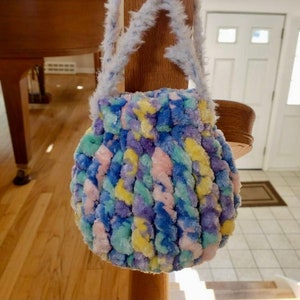 Peut inclure: Un petit sac rond en crochet fait de fil pelucheux aux couleurs pastel. Le sac a une bandoulière blanche et moelleuse.