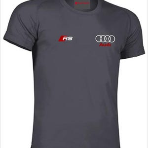 Peut inclure: T-shirt gris foncé avec le logo Audi et les lettres "RS" en rouge et blanc. Le logo Audi est composé de quatre anneaux entrelacés. Le t-shirt a des manches courtes et un col rond.