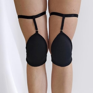 Può includere: Ginocchiere nere con cinghie regolabili. I cuscinetti a forma di goccia sono attaccati a una fascia nera che avvolge la coscia. I cuscinetti sono collegati alla fascia della coscia con una sottile cinghia e hardware color oro.