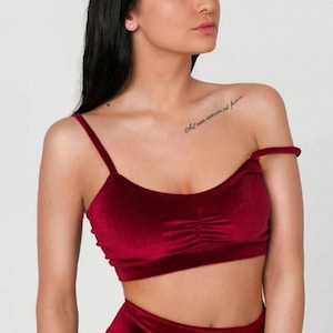 May include: A red velvet crop top with thin straps. The top has a gathered detail in the front. The model is wearing the top with a matching skirt. The text "Oh I am invincible but fearless" is tattooed on the model's shoulder.