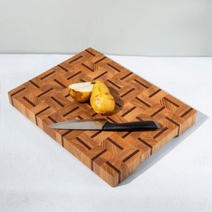 Könnte beinhalten: Ein hölzernes Schneidebrett mit geometrischem Muster, einem Edelstahlmesser und zwei Birnen. Das Brett besteht aus hellem und dunklem Holz, und die Birnen sind gelb. Das Messer hat einen schwarzen Griff.