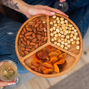 May include: A wooden serving tray with three sections, filled with almonds, dried apricots, and hazelnuts. The tray is round and has a natural wood finish.