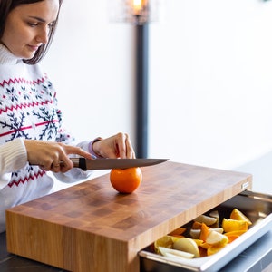 May include: A wooden cutting board with a built-in drawer for storing produce. The drawer is open and contains sliced oranges, lemons, and limes. A person is using a knife to cut an orange on the board.
