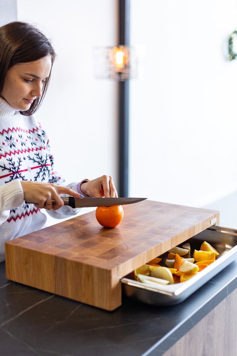 May include: A wooden cutting board with a built-in drawer for storing produce. The drawer is open and contains sliced oranges, lemons, and limes. A person is using a knife to cut an orange on the board.