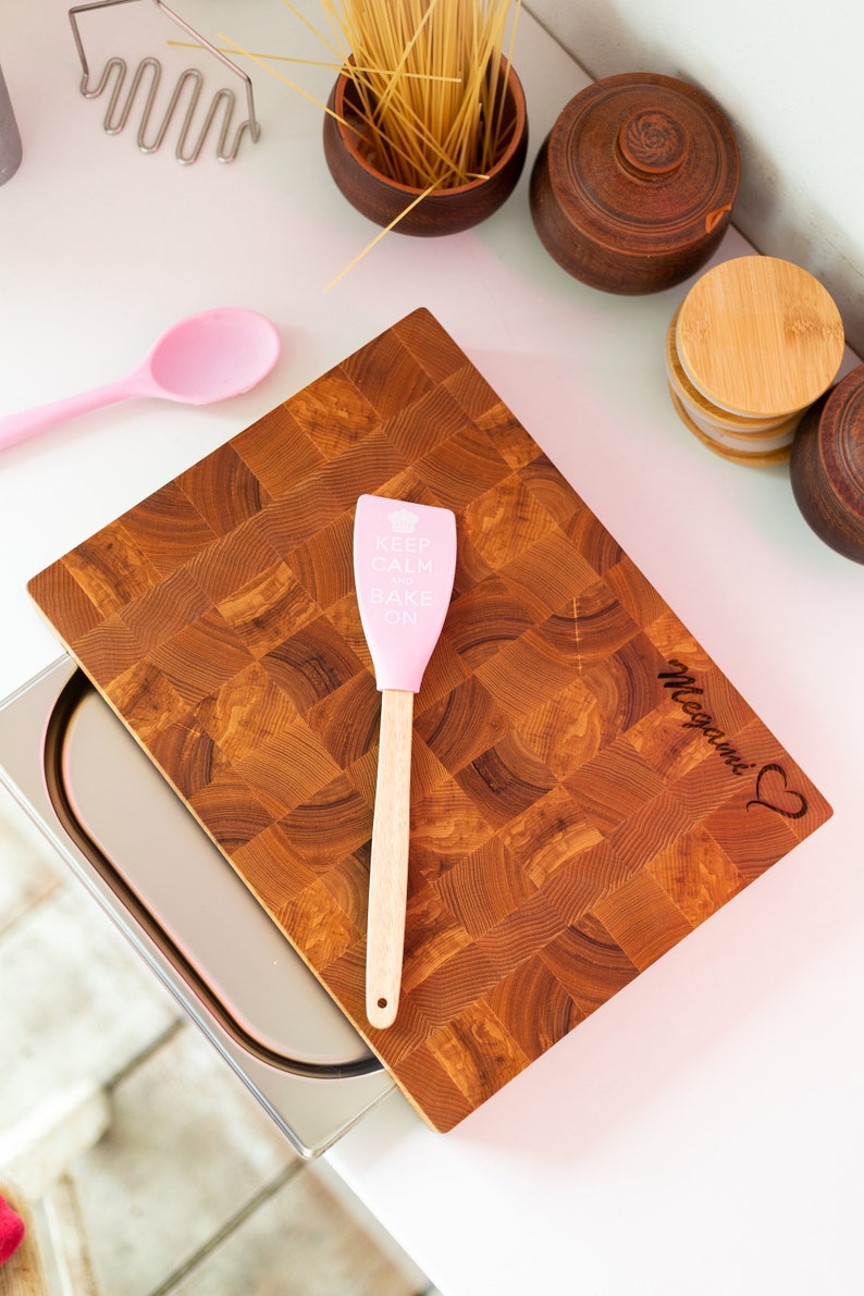 May include: A wooden cutting board with a pink spatula that says "Keep Calm and Bake On". The board is engraved with the name "Megan" and a heart.