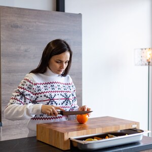 May include: A woman wearing a white sweater with a red and black snowflake pattern is cutting an orange fruit on a wooden cutting board with a built-in drawer. The drawer is open and contains a silver tray with fruit inside.
