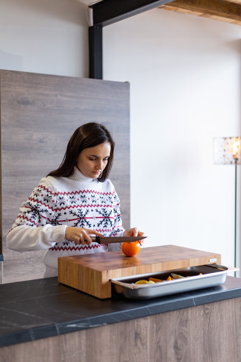 May include: A woman wearing a white sweater with a red and black snowflake pattern is cutting an orange fruit on a wooden cutting board with a built-in drawer. The drawer is open and contains a silver tray with fruit inside.