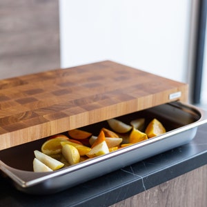 May include: A wooden cutting board with a checkered pattern sits on top of a stainless steel baking dish filled with sliced fruit. The fruit is a mix of yellow and orange, likely pears and oranges.