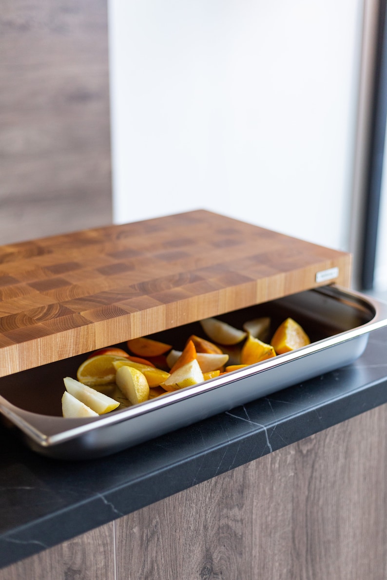 May include: A wooden cutting board with a checkered pattern sits on top of a stainless steel baking dish filled with sliced fruit. The fruit is a mix of yellow and orange, likely pears and oranges.