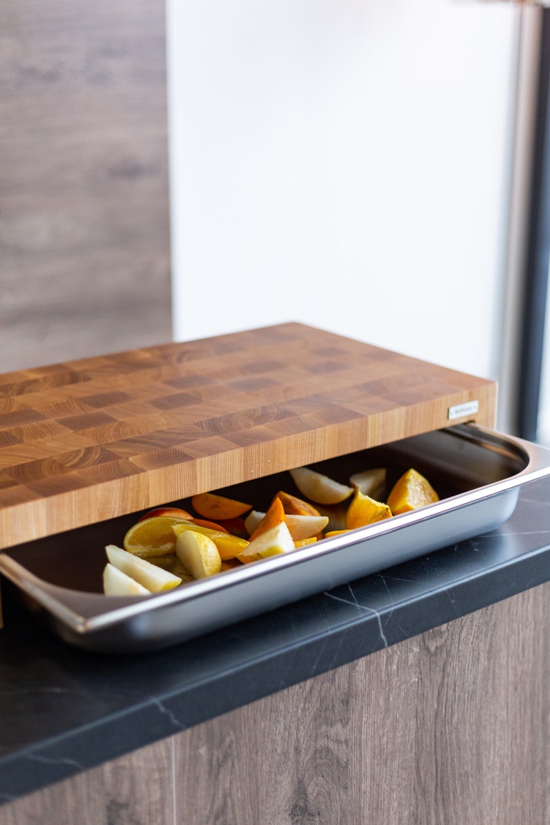 May include: A wooden cutting board with a built-in stainless steel tray. The tray is filled with sliced fruit, including pears, oranges, and persimmons.