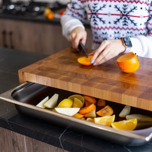 May include: A wooden cutting board with a built-in drawer, filled with sliced oranges and persimmons. A person is cutting a persimmon on the board.