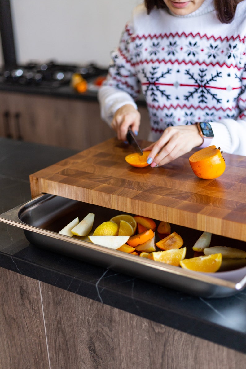 May include: A wooden cutting board with a built-in drawer, filled with sliced oranges and persimmons. A person is cutting a persimmon on the board.