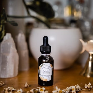 May include: A dark brown glass bottle with a black dropper top and a silver charm. The bottle is labeled "Moon Glow Botanicals Grounding & Centering Oil" and has a white background with black text. The bottle is sitting on a wooden surface with dried flowers scattered around it.