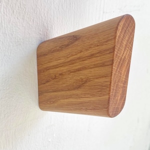 Puede incluir: Un gancho de pared de madera con un borde redondeado. El gancho está hecho de madera marrón claro con un patrón de veta natural.