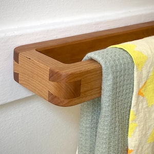 May include: Wooden towel rack mounted on a white wall. The rack is made of light brown wood with visible joinery. Two towels are draped over the rack: a light green waffle-weave towel and a cream-colored towel with yellow floral patterns.