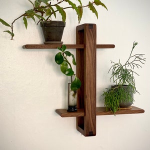 May include: A wooden wall shelf with three shelves, each holding a potted plant. The shelf is made of dark wood and has a simple, modern design.