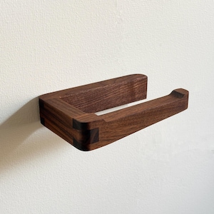 May include: A wooden toilet paper holder mounted on a white wall. The holder is made of dark brown wood with visible grain and features a rectangular design with rounded edges. The holder is designed to hold a single roll of toilet paper.