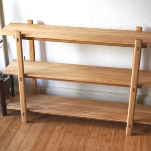 May include: A three-tiered wooden shelf unit. The shelves are made of light-colored wood and are supported by vertical posts. The unit is approximately 48 inches wide, 36 inches tall, and 12 inches deep. The design is simple and modern.
