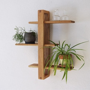 May include: A wooden wall shelf with four shelves, featuring a geometric design. The shelf is made of light brown wood and has a natural finish. Two small glass vases and two potted plants are displayed on the shelves.