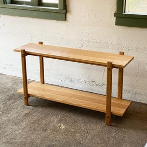 May include: A two-tiered wooden shelf unit with a natural wood finish. The shelf is made of solid wood and has a simple, modern design.