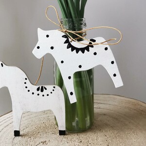 May include: Two white wooden horses with black painted details, including polka dots and a geometric pattern, are displayed on a wooden surface. The horses are tied together with twine and are positioned in front of a glass vase filled with green stems.