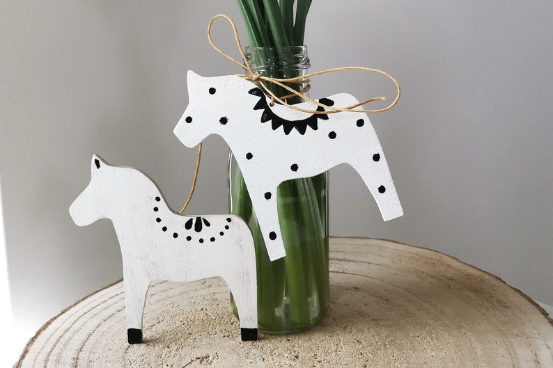 May include: Two white wooden horses with black painted details, including polka dots and a geometric pattern, are displayed on a wooden surface. The horses are tied together with twine and are positioned in front of a glass vase filled with green stems.