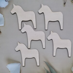 May include: Five wooden cutout horses in the shape of a traditional Swedish Dala horse. The horses are all white and are arranged in a semi-circle on a light brown background.