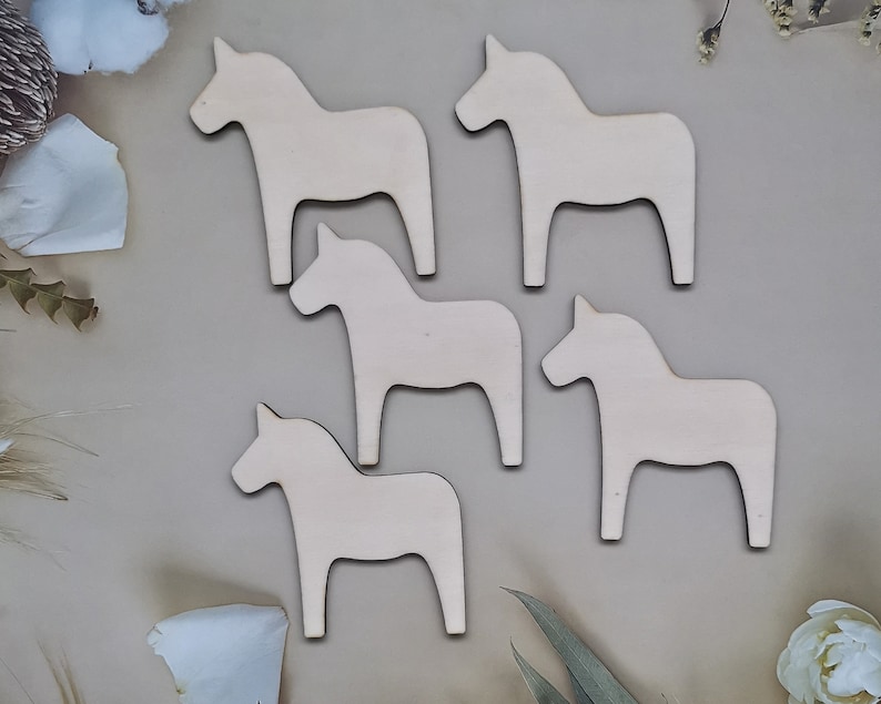 May include: Five wooden cutout horses in the shape of a traditional Swedish Dala horse. The horses are all white and are arranged in a semi-circle on a light brown background.