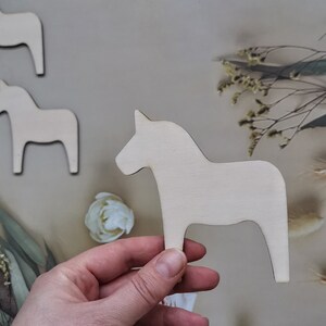 May include: A set of four unfinished wooden horse figurines. The horses are all the same size and shape, and they are standing on a light brown surface.