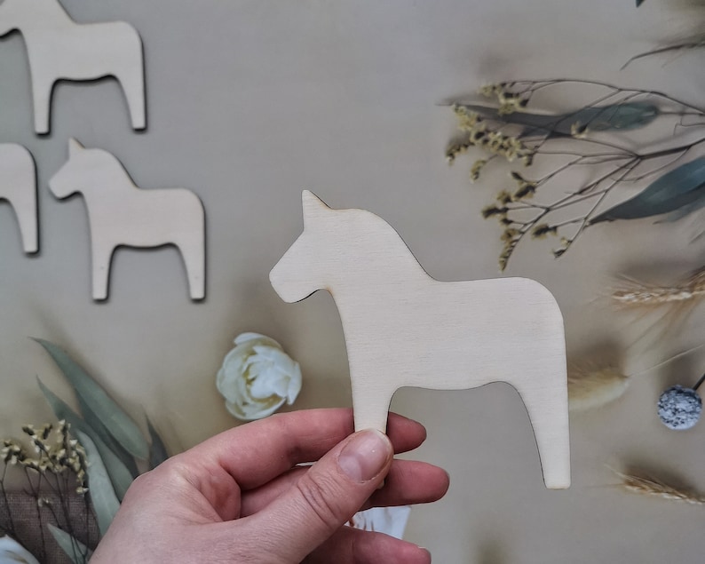May include: A set of four unfinished wooden horse figurines. The horses are all the same size and shape, and they are standing on a light brown surface.