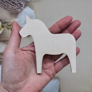 May include: A white wooden Dala horse figurine, a traditional Swedish folk art piece, is held in a person's hand.