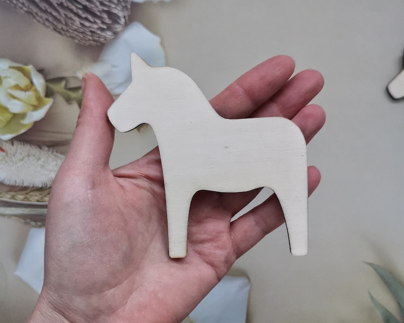 May include: A white wooden Dala horse figurine, a traditional Swedish folk art piece, is held in a person's hand.