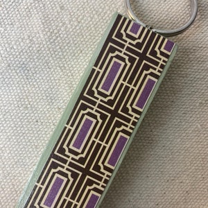 Puede incluir: Un llavero rectangular verde y marrón con un patrón geométrico con acentos morados. El llavero tiene un anillo de metal plateado.